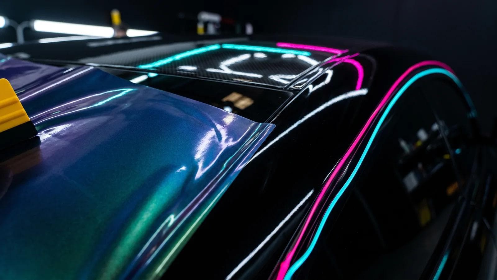 Ausschnitt einer glänzend schwarzen Sportwagenflanke, zur Hälfte mit einer neuen Farbfolie beklebt und mit angedeutetem Rakel, in türkis-magentafarbenem Neonlicht.