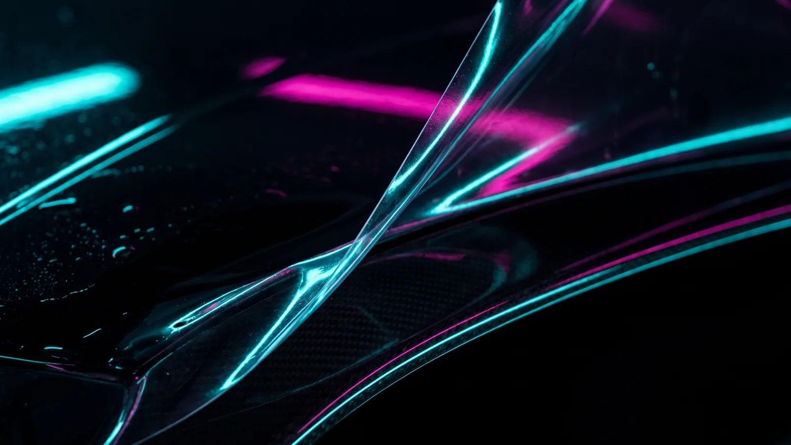 Nahaufnahme einer glasklaren PPF-Lackschutzfolie, die an der steinschlaggefährdeten Kante einer schwarzen Motorhaube angelegt wird, in türkis-magentafarbenem Neonlicht.